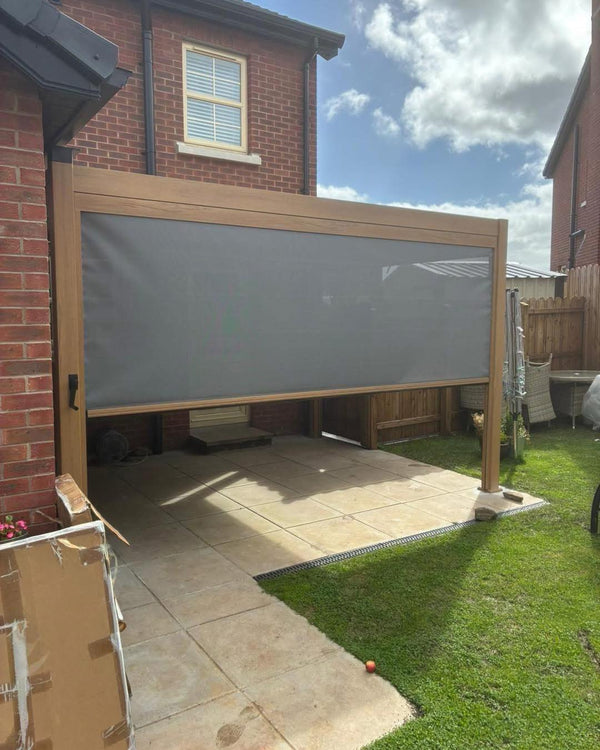 Pergola installed by Jacksons of Saintfield in Northern Ireland. Outdoor patio area with a large screen covering the space, set against a brick house and blue sky.